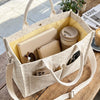 Un sac en velours beige contenant un portefeuille rose pâle, une tasse en céramique et un smartphone sur une table en bois.