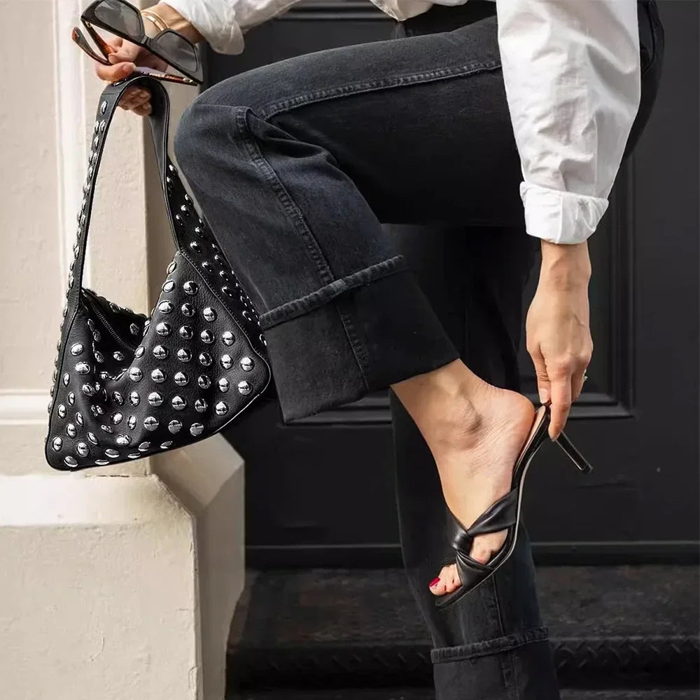 A person wearing dark jeans and a white shirt holds a black designer metal rivet shoulder bag.