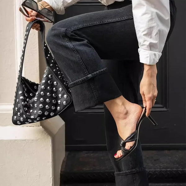 A person wearing dark jeans and a white shirt holds a black designer metal rivet shoulder bag.