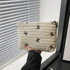 A beige quilted makeup pouch with black embroidered leaves is held against a white window blind.