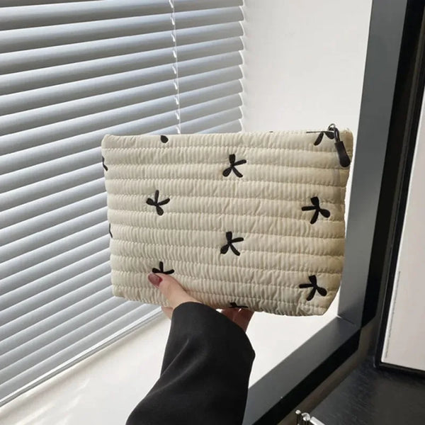 Pochette de maquillage beige matelassée avec des feuilles noires brodées, tenue contre un store blanc.