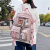 A woman wearing a white sweater carries a pink Korean style large capacity backpack with clear compartments and a white pom-p