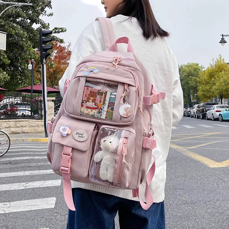 A woman wearing a white sweater carries a pink Korean style large capacity backpack with clear compartments and a white pom-p