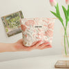 A hand holds a pink floral canvas makeup bag with embroidered details next to a vase of tulips.