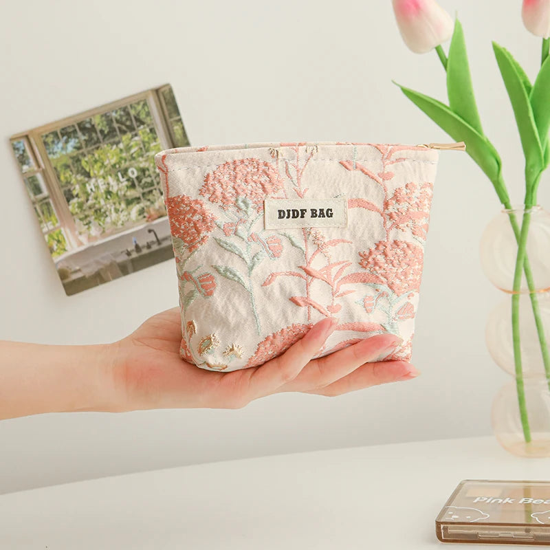 A hand holds a pink floral canvas makeup bag with embroidered details next to a vase of tulips.
