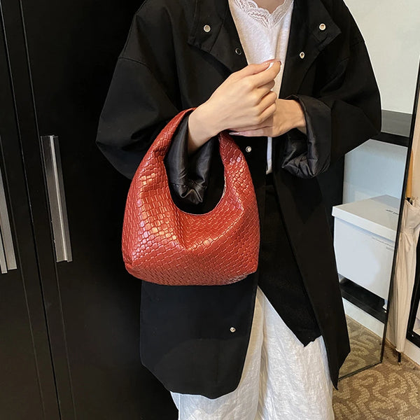 A person holds a reddish-brown woven embossed hobo shoulder bag with a top handle against a black jacket and white shirt.
