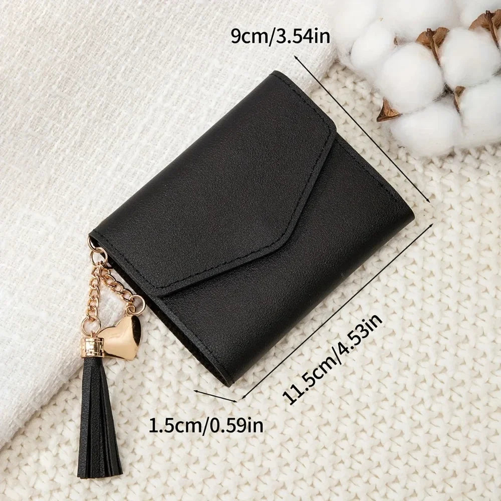 Black PU leather coin clutch wallet with a gold heart charm and black tassel resting on a cream textured fabric.