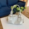 A white PU leather shoulder bag with silver rivets and a handle is displayed on a white table next to a blue sofa.