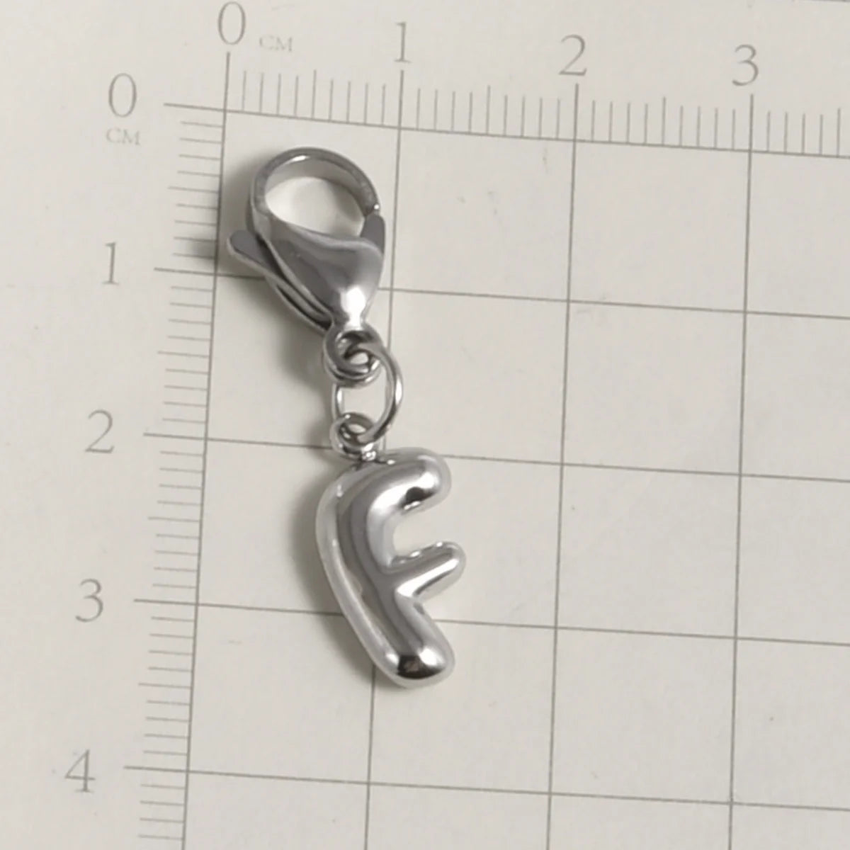 Silver stainless steel alphabet letter charm with a lobster clasp placed on a ruler.