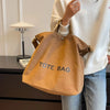 A person holds a tan canvas tote bag with blue lettering in front of a dark surface.