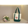 A white canvas tote bag with green straps and a red logo hanging against a light gray wall.