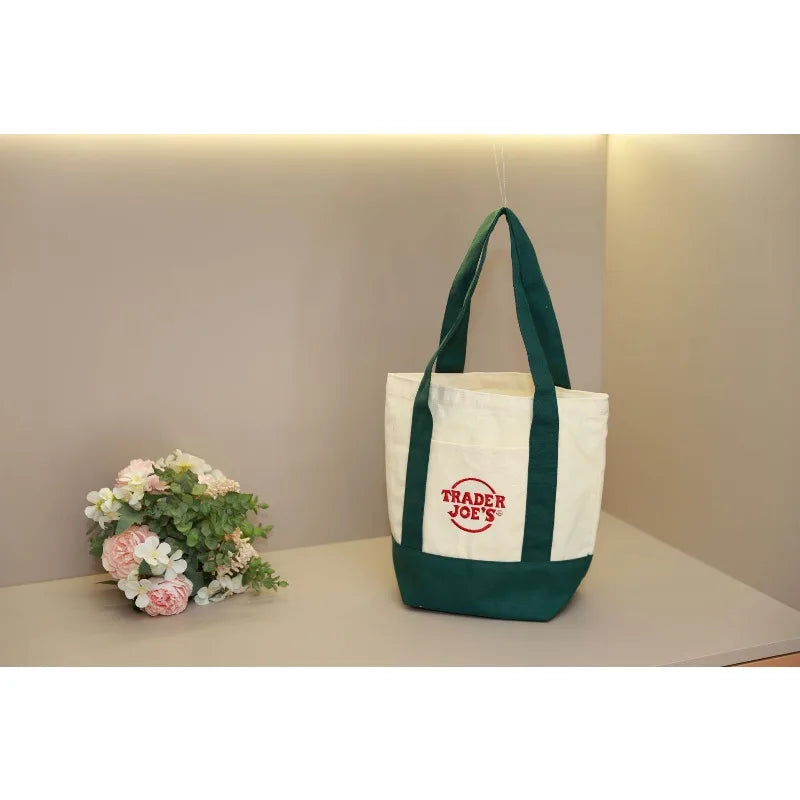 A white canvas tote bag with green straps and a red logo hanging against a light gray wall.