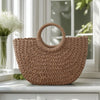 A brown handwoven rattan beach bag resting on a white surface near a window.