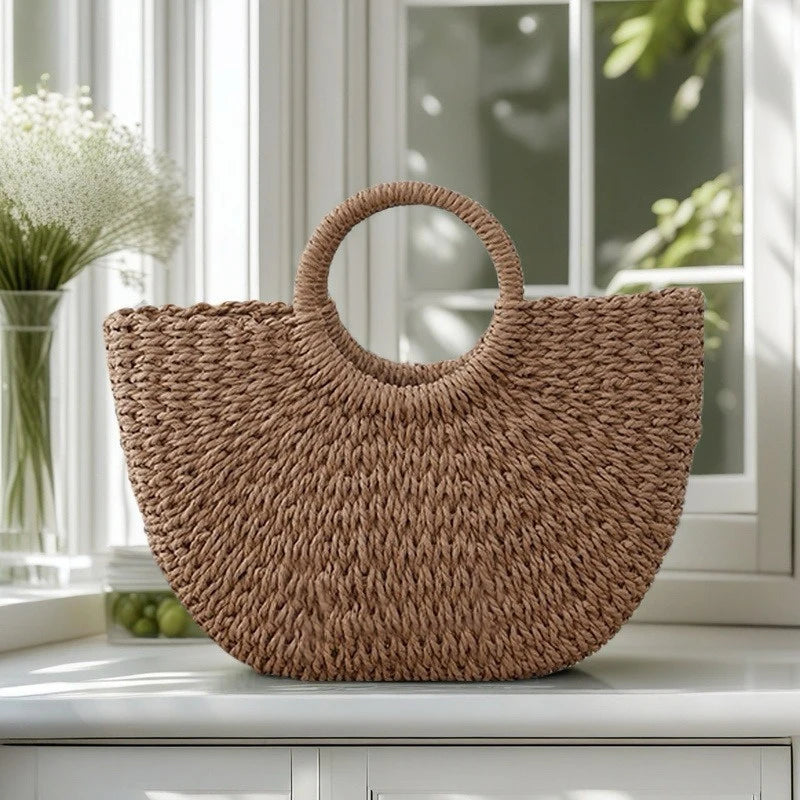 A brown handwoven rattan beach bag resting on a white surface near a window.