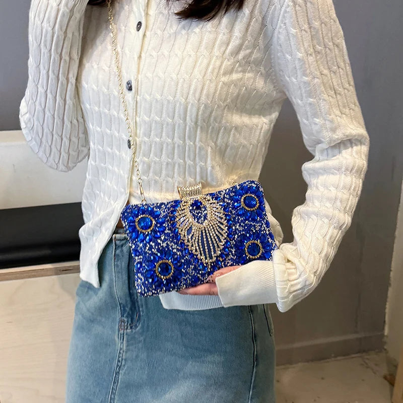 A white knitted sweater sleeve shows a blue rhinestone evening clutch bag with gold accents.