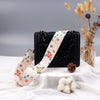 Black crossbody bag with a light floral pattern strap resting on white fabric next to dried flowers and a pine cone.