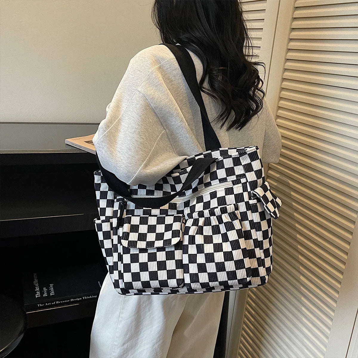 A woman is holding a black and white checkered corduroy tote bag against a light-colored vertical blind.