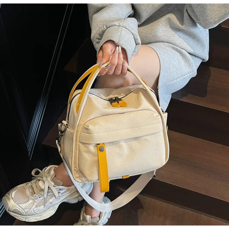 A light beige nylon crossbody bag with a yellow top handle is being held by a person on wooden stairs.