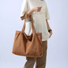 A woman holds a large brown canvas shoulder tote bag and wears a white t-shirt and tan pants.