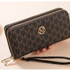 A dark brown patterned wallet with gold zipper details and a black strap is shown being held by a hand.
