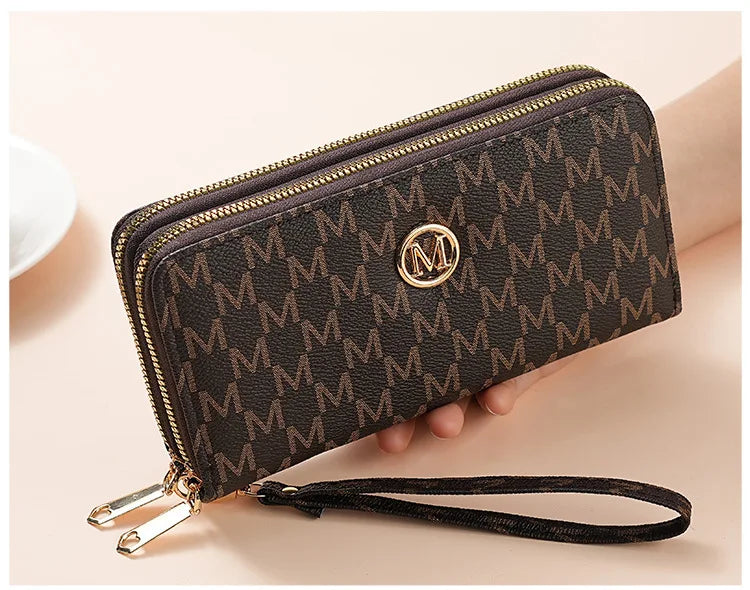 A dark brown patterned wallet with gold zipper details and a black strap is shown being held by a hand.