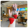 Une main tient un porte-clés en forme de sac de fraises rouge vif avec une sangle en cuir, sur un fond intérieur flou.
