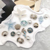 A collection of golden and iridescent sea-themed shoe charms including starfish, conches, and pearls displayed on a white sur