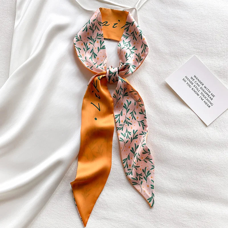 Foulard de cheveux floral en soie orange noué en nœud, posé sur un tissu blanc avec une petite carte.