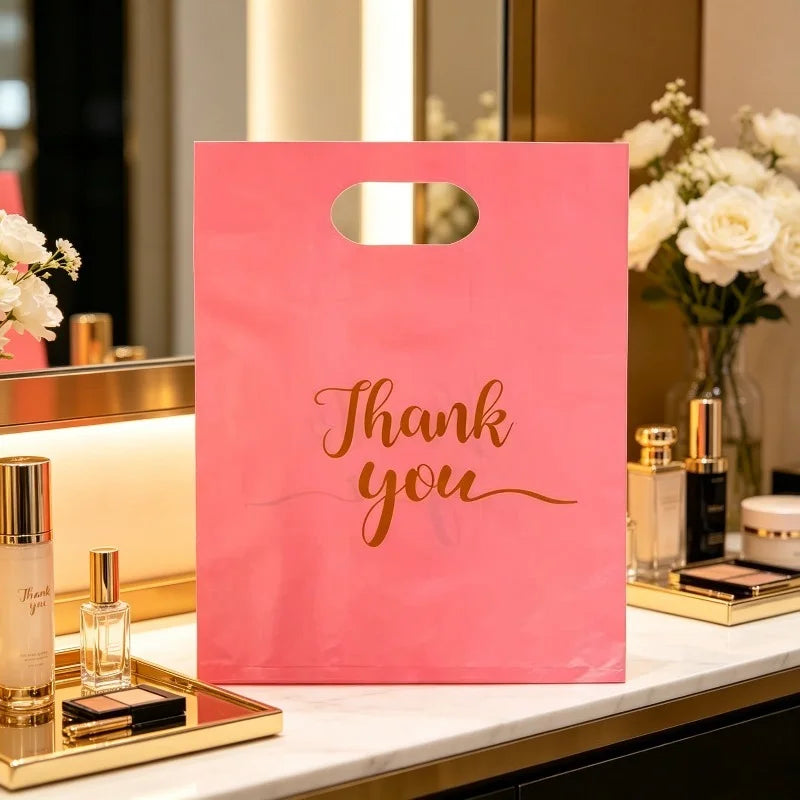 A pink bulk thank you gift bag with gold script text resting on a white marble surface next to perfume bottles.