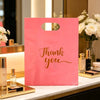 A pink bulk thank you gift bag with gold script text resting on a white marble surface next to perfume bottles.