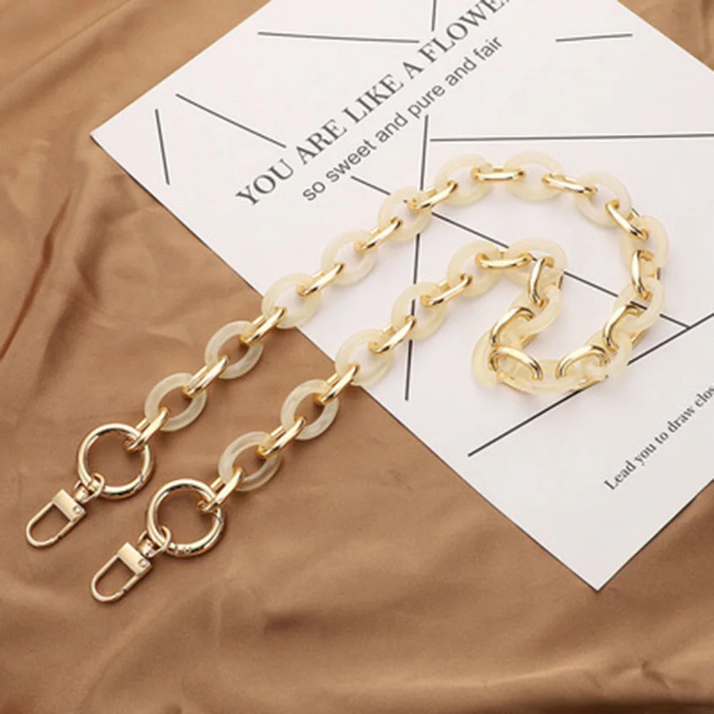 A gold-toned acrylic handbag strap chain is displayed on a piece of paper against a brown fabric background.