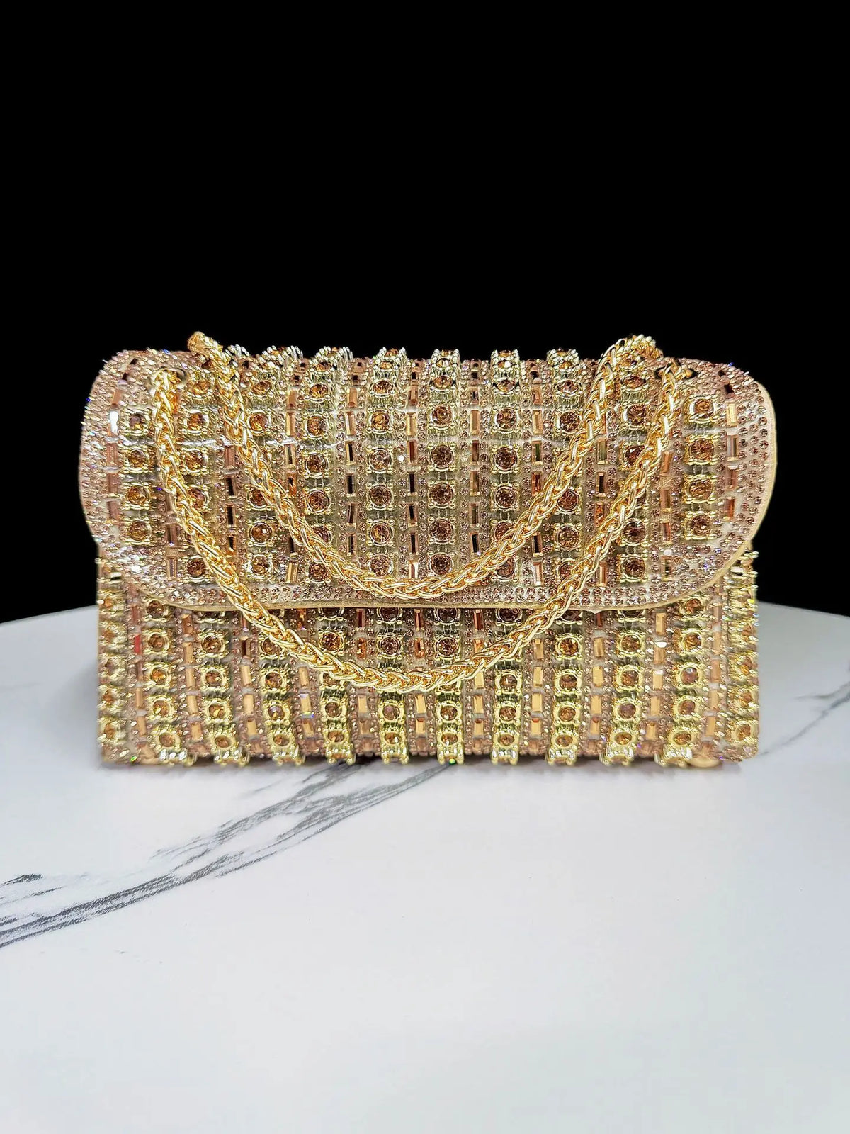 A rectangular gold and rose gold rhinestone evening clutch bag with a diamond chain resting on a white marble surface.
