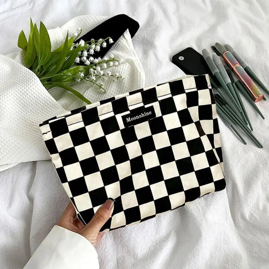 A black and white checkered cosmetic storage bag labeled Moonshine is shown next to green foliage and makeup brushes on a whi