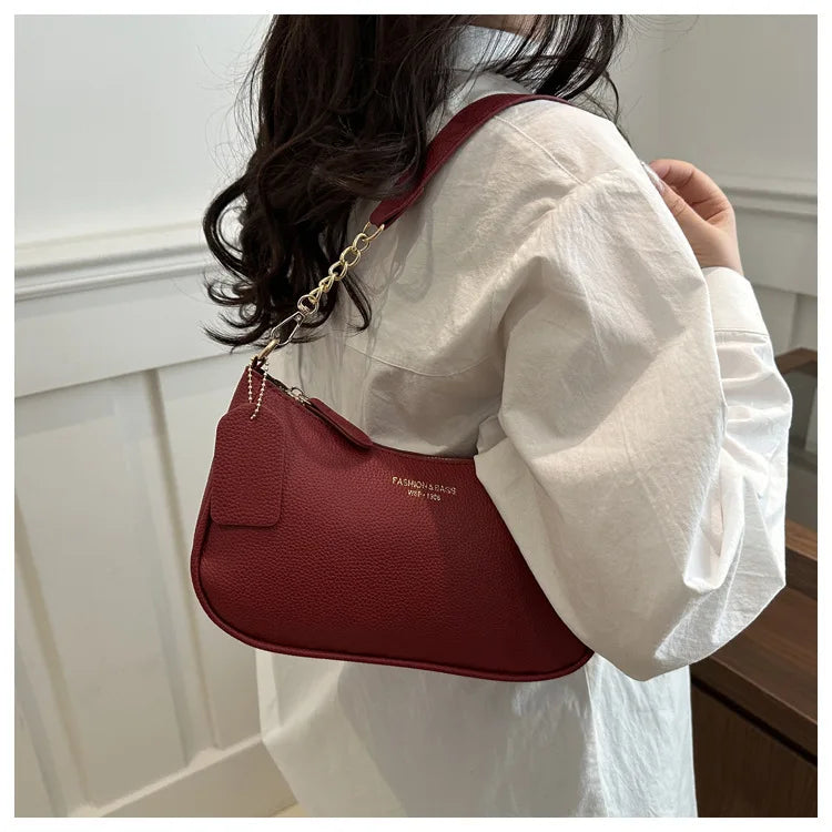 A woman holds a maroon leather shoulder bag with a gold chain pendant against a white shirt.