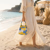 A woman wearing a long cream lace dress holds a yellow and blue geometric colorblock knitted bucket bag on a beach.