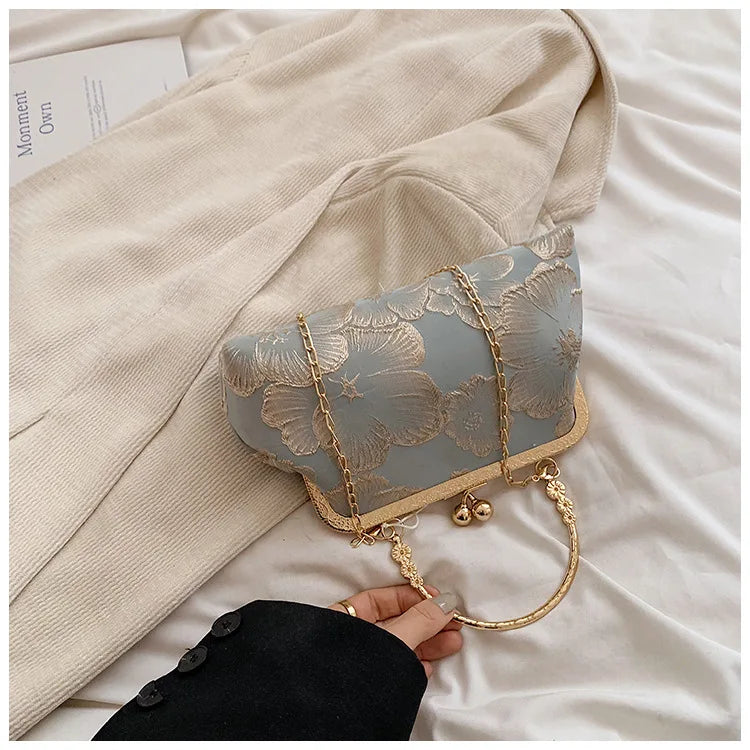 A light blue embroidered silk evening clutch with a gold chain strap resting on a beige fabric.