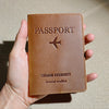 A brown leather passport holder with a stitched design and a small airplane emblem is held in a hand.
