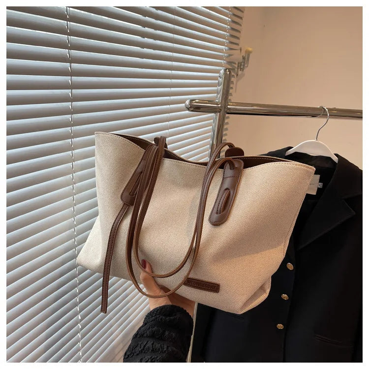 A beige canvas shoulder tote bag is displayed hanging on a dark jacket next to a window blind.