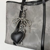 A gray faux leather tote bag with a black heart charm and silver beaded key chain hanging from the strap.