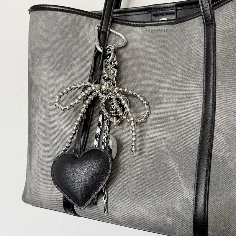 A gray faux leather tote bag with a black heart charm and silver beaded key chain hanging from the strap.