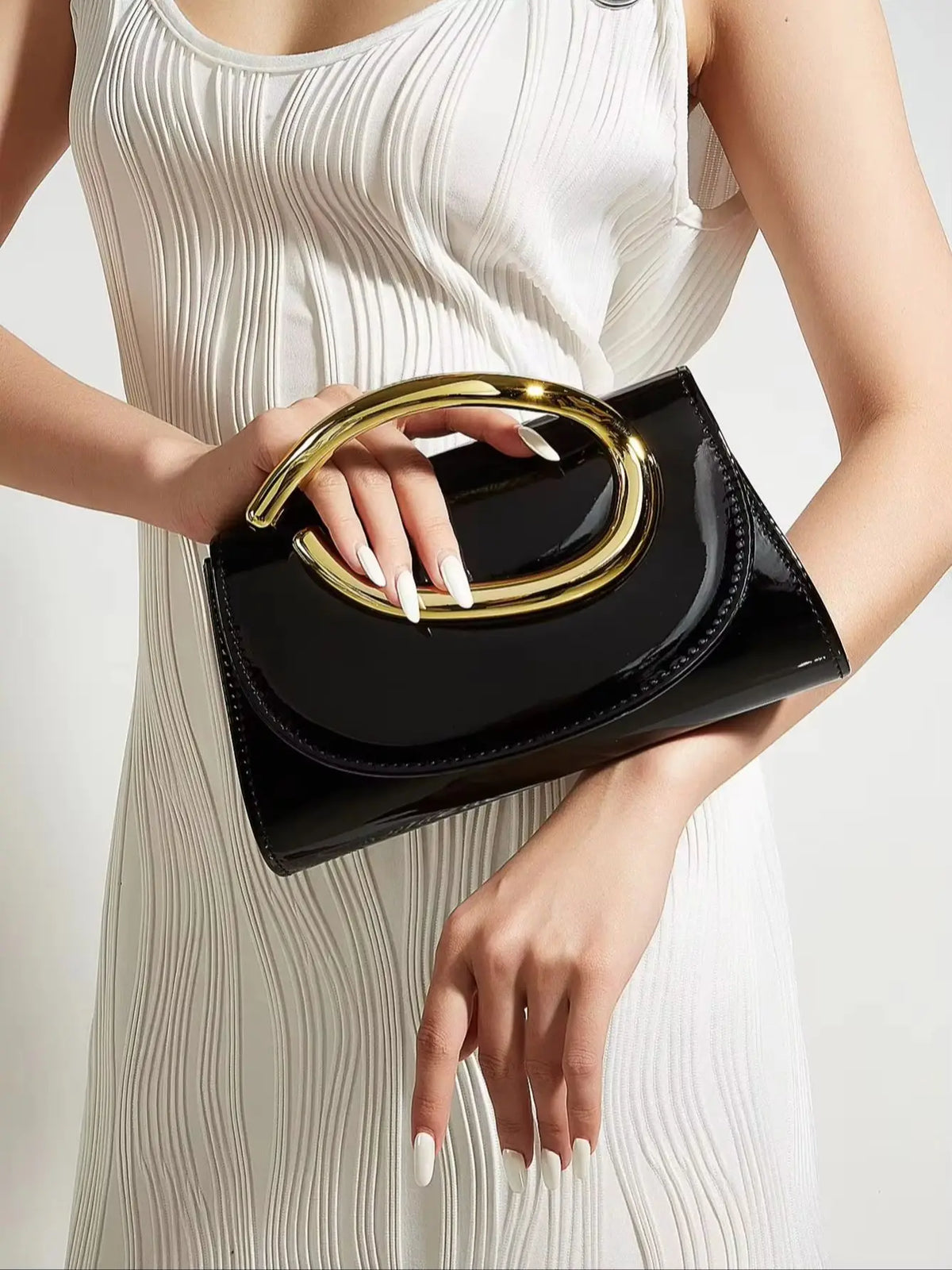 A woman holds a black clutch purse with a gold handle against a white ribbed dress.
