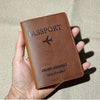 A brown leather passport holder and travel wallet with a stitched logo is held in a hand against a beige fabric background.