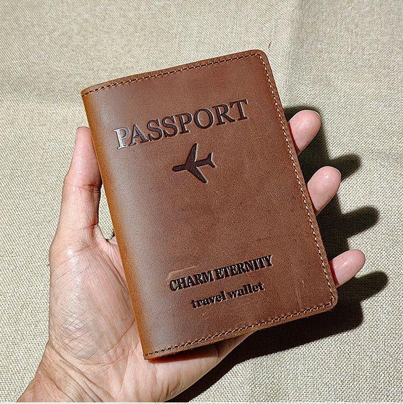 A brown leather passport holder and travel wallet with a stitched logo is held in a hand against a beige fabric background.