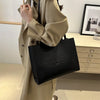 A person wearing a beige trench coat holds a black tote bag in front of a light wood cabinet.