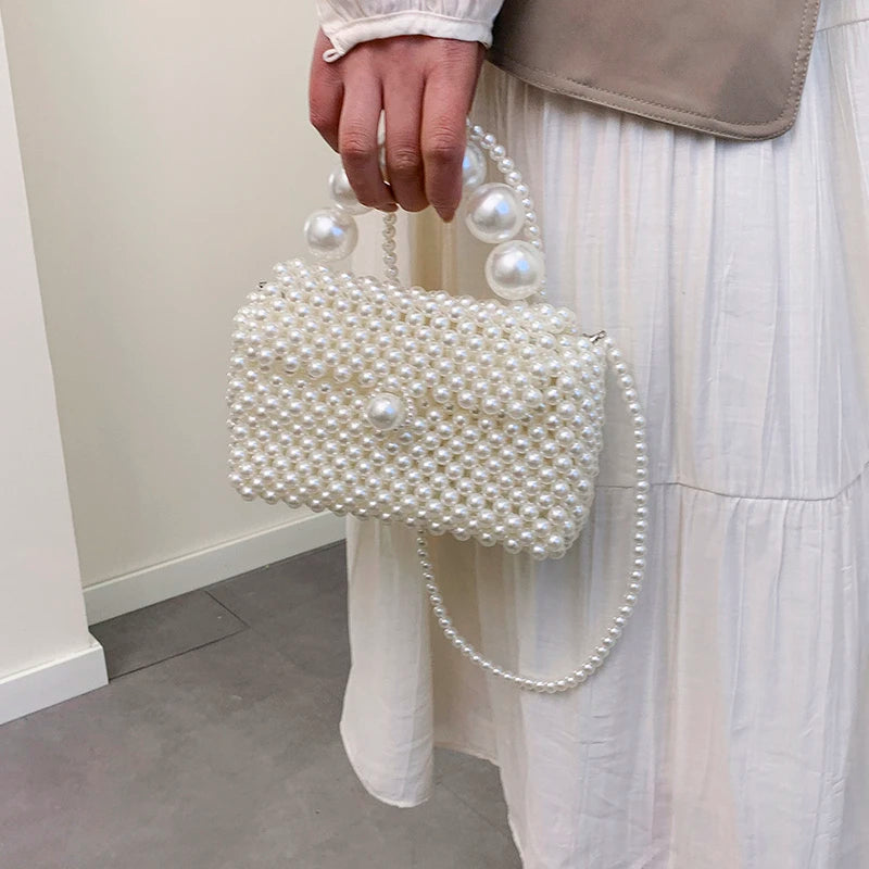 A hand holds a white pearl beaded clutch bag with a pearl strap against a white dress.