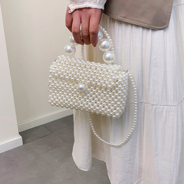 A hand holds a white pearl beaded clutch bag with a pearl strap against a white dress.