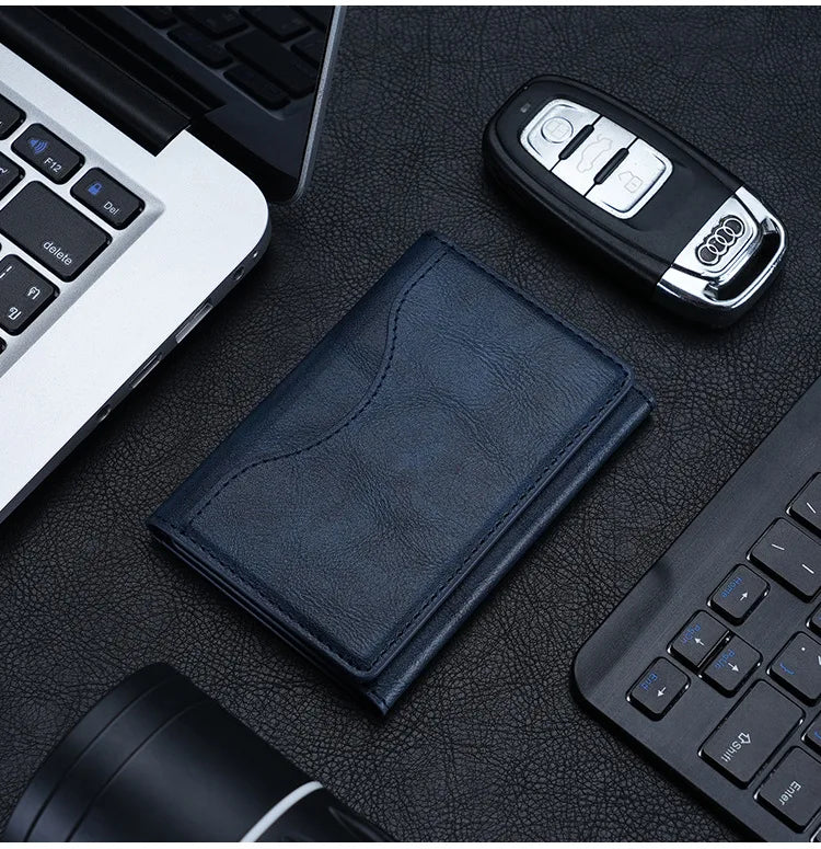 A dark blue magnetic leather card wallet resting on a black surface next to a laptop and a stylus.