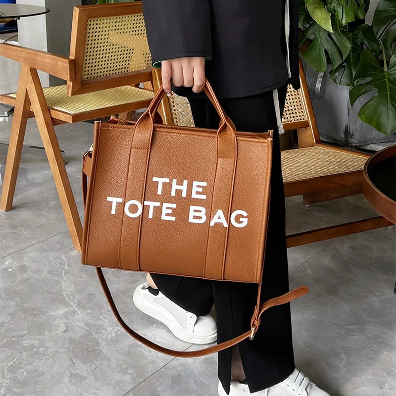 Un sac fourre-tout en cuir marron avec du texte blanc est tenu par une personne debout à côté de chaises en bois.