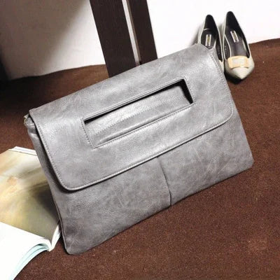 A gray leather crossbody clutch bag resting on a brown surface with a pair of black dress shoes nearby.