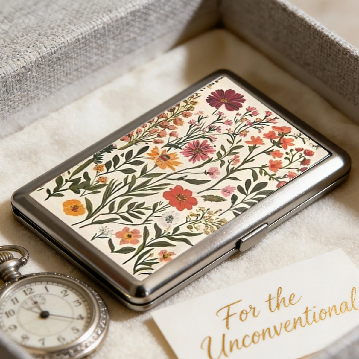 An open metal card case with a floral pattern resting on a light fabric background.